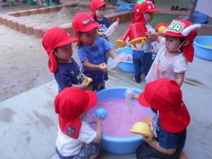水色のたらいに入ったピンク色の水をマヨネーズの空容器やカップで水をすくって遊んでいる赤い帽子を被った子ども達の写真