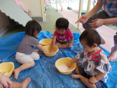 ビニールシートの上に座っているさな子ども達が黄色の洗面器の中に手を入れて遊んでいる写真