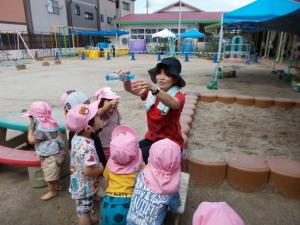 外の砂場でジュース屋さんをする先生と見ている子供達の写真