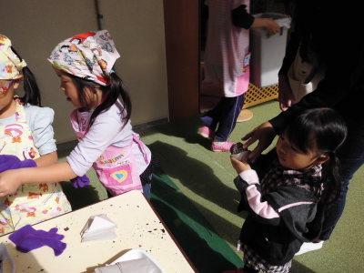 焼き芋を手に持つ小さな女の子と横の机の後ろで話をしている園児2名の写真