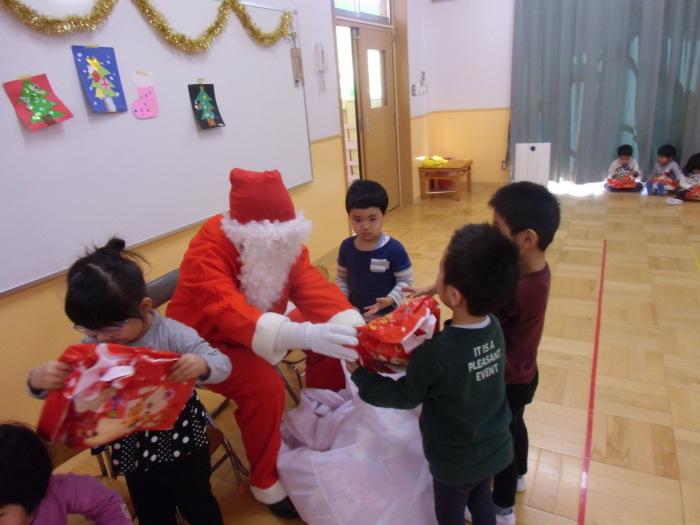 教室の椅子に赤い洋服を着たサンタクロースが座っており、大きな白い袋から取り出したプレゼントを子ども達がもらっている写真