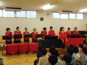 赤い衣装で揃えたトーンチャイム演奏の方たちが横一列に並んで立っており、子ども達が椅子に座って話を聞いている写真