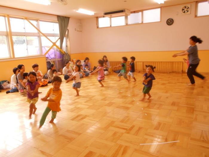 保育所の園児達が走り回りながらリズム遊びを披露し、それを見ているたくさんの親子の写真