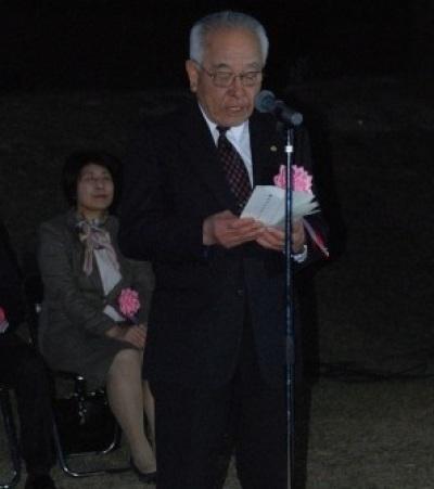 胸に桃色の胸章を付けてスーツを着た白髪の男性がスタンドマイクの前に立って話をしている写真