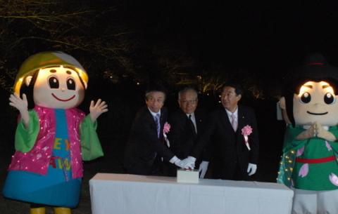 はちかづきちゃんとねや丸くんが両脇に立っており、設置されている机の上に置かれたボタンに3人の男性が手を重ねてライト点灯のボタンを押している写真