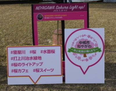 撮影用インスタボードが置かれ、「＃寝屋川 ＃桜 ＃水面桜 ＃打上川治水緑地 ＃桜のライトアップ ＃桜カフェ ＃桜スイーツ」「中核市ねやがわ～食と笑顔が輝くまち～」と 吹き出しに描かれたプレートが立て掛けられている写真