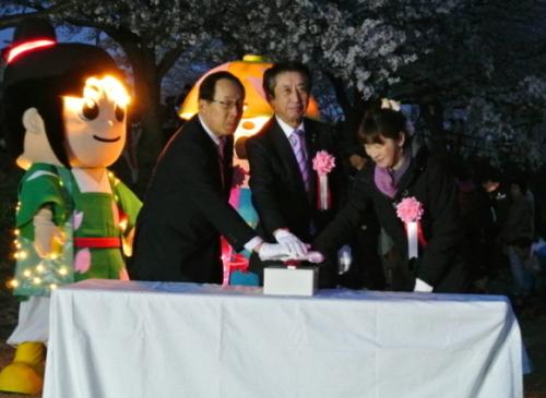 電気で光っているはちかづきちゃんの横で市長、関係者の男女1名づつで点灯スイッチに片手をおいている写真