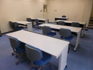青い絨毯が敷かれており、2人ずつ座れる机が2列にならんでいるITセミナー室の写真