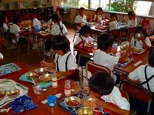 各班に分かれた制服姿の子供たちが机を並べて、給食を食べている様子を教室斜めから写した写真
