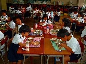 各班に分かれた制服姿の子供たちが机を並べて、給食を食べている様子を教室前方から写した写真