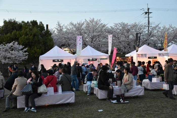 出店しているお店の白いテントが並び、飲食スペースに設けられている席に座って、沢山のお客さんで賑わっている桜カフェの様子の写真