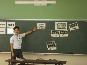 黒板に貼られた用紙を指さしている男性教員の写真