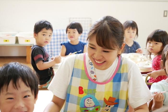 笑顔で子供たちに接しているかわいいエプロン姿の倉内さんと周りで給食を食べている園児の写真