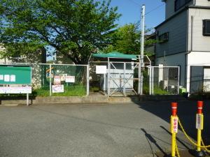 堀溝小学校の校庭の樹木の前に緑色の屋根が設置してある場所に水質自動監視装置が設置してあり、機械の前は金網の柵がしてある写真