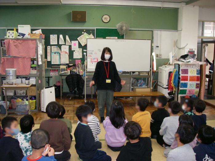 名札を首からぶら下げた職員の女性が子ども達に話をしている学童保育での教室の様子の写真
