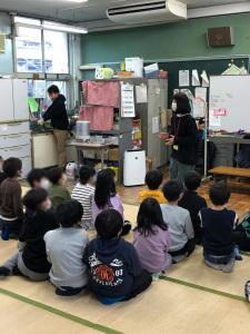 教室の中で子ども達がござの上に座って学童保育の先生の話を聞いている写真