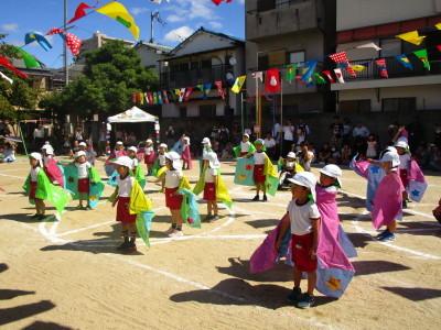万国旗の飾られた園庭で白い帽子に色とりどりのマントを身にまとった子供たちが団体演技をしている写真